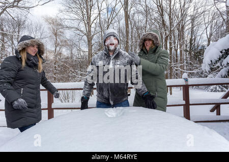 Due Yong donne In cappotti invernali scherzosamente spingere un giovane Mans faccia nella neve Foto Stock