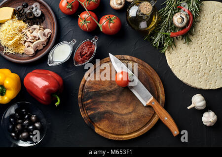 Il taglio di legno con pizza tradizionale preparazione ingredienti: formaggio, pomodori, salsa, olive, olio d'oliva, pepe, spezie. Nero texture di sfondo della tabella Foto Stock