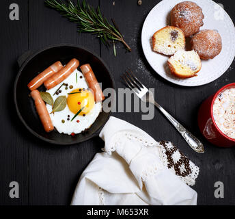 Uova fritte con salsicce in un nero padella, cacao con marshmallows e muffin su tavoli in legno nero, vista dall'alto Foto Stock