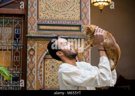 Giovani Musulmani uomo in abbigliamento tradizionale tenendo un gatto giallo in un tradizionale ambiente Marocchino davanti a una parete decorata Foto Stock