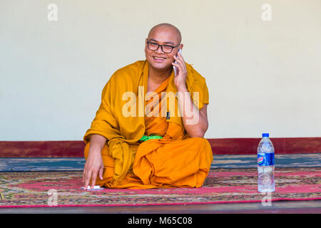 Sorridente monaco buddista sul telefono cellulare mentre seduto sul pavimento con una bottiglia di acqua a Shwedagon pagoda Yangon, Myanmar (Birmania), l'Asia in febbraio Foto Stock