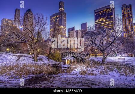 Scena invernale di Gapstow Bridge in Central Park di NY illuminata con alto-aumento in background. Le aree circostanti e alberi coperte con il bianco della neve. Foto Stock