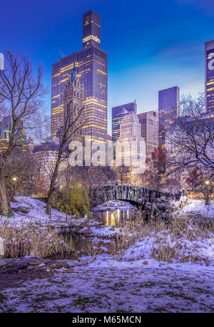 Scena invernale di Gapstow Bridge in Central Park di NY illuminata con alto-aumento in background. Le aree circostanti e alberi coperte con il bianco della neve. Foto Stock