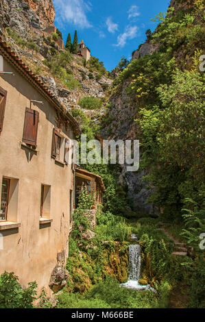 Vista delle case vicino al torrente e bluff con chiesa su scogliere nell'affascinante Moustiers-Sainte-Marie village. Nella regione della Provenza, Francia sudorientale. Foto Stock