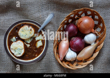 La zuppa di cipolla con blue stilton formaggio gruviera con aglio, Echalion Francese di scalogni e rosso e marrone e le cipolle in un cesto di vimini su un panno di Hesse backg Foto Stock