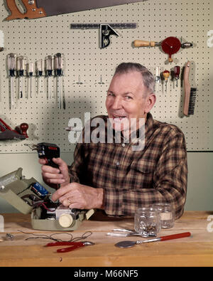 Anni sessanta anni settanta sorridente uomo SENIOR HOLDING UTENSILE SALDATORE A IN CASA O GARAGE NEGOZIO guardando la fotocamera - KS3179 HAR001 HARS in interni che fissa la nostalgia di mezza età uomo di mezza età sparso il contatto visivo 45-50 anni 50-55 anni CHORE HOBBY HOBBY RIPARAZIONE FIX WORKBENCH persone maschi adulti la saldatura SALDATORE ETNIA CAUCASICA MONTARLO FISSARLO guardando la telecamera MISTER VECCHIO tabellone persone pistola per saldatura tettoia dell'attrezzo Foto Stock