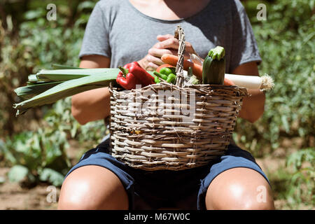 Primo piano di un giovane uomo caucasico con un rustico cesto pieno di ortaggi appena raccolti in un frutteto organico Foto Stock