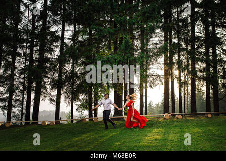 Bella coppia felice in amore è in esecuzione per mano lungo il verde foresta estate durante il tramonto. Foto Stock