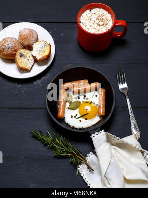 Uovo fritto con salsicce in un nero padella, cacao con marshmallows e muffin su tavoli in legno nero, vista dall'alto Foto Stock