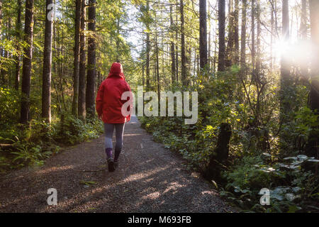 Ragazza che indossa un luminoso giacca rossa è a piedi i bellissimi boschi durante una vibrante inverno mattina. Preso in Ucluelet, Isola di Vancouver, BC, Canada. Foto Stock