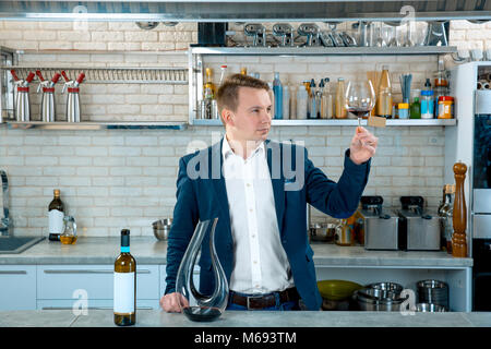 Concentrato giovane sommelier degustazione e guardando il vino rosso in vetro su cucina interno Foto Stock