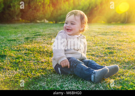 Carino baby boy all'aperto, piccola e dolce bambino seduto sul verde e fresco campo di erba in una luminosa giornata di sole, godendo della prima molla giorni felici childhoo spensierata Foto Stock