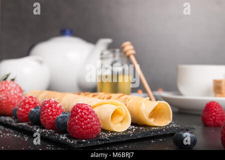 Frittelle di laminati con fresche di fragola e lampone e mirtillo su una pietra nera piastra al buio su un tavolo di pietra con il miele e lo zucchero bianco e co Foto Stock