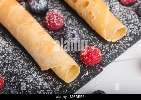 Frittelle di laminati con fresche di fragola e lampone e mirtillo su una pietra nera piastra su un bianco tavolo in legno con il miele e lo zucchero bianco e Foto Stock