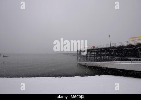 Molo di Southend innevato e spiaggia con nuvole di neve. Nevicando. Capanne colorate. Gambe di ferro. Barca Foto Stock