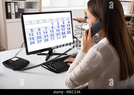 Close-up di un giovane imprenditrice parlando al telefono di rete fissa con il Calendario sullo schermo del computer Foto Stock