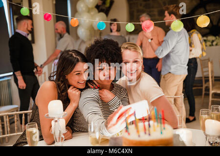Tre donne siedono ad un tavolo a una festa di compleanno. Si sta utilizzando un telefono intelligente per prendere un divertente selfie. Foto Stock