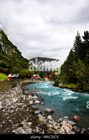 Red scandinavian case di legno e cabine a un selvaggio fiume turchese e il litorale circondato da montagne e di fitta foresta e gli alberi in Norvegia Foto Stock