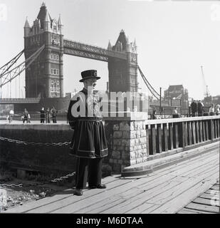 Degli anni Cinquanta, foto storiche di un Yeoman Warder in 'abito quotidiano' uniforme al di fuori in piedi la Torre di Londra, con il fiume Tamigi e il Tower Bridge dietro di lui. Ex-veterano, essi svolgono un ruolo importante come custodi di cerimoniale presso la torre e sono popolarmente noto come Beefeaters. Foto Stock