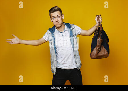 Elegante bionda giovane uomo in nero jeans e t-shirt bianco, è in piedi con la borsa nella sua mano isolata a sfondo giallo. Foto Stock