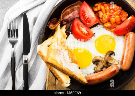 Prima colazione inglese tradizionale - pancetta, salsicce, uova fritte, pomodori, fagioli, funghi, toast con burro - nel recipiente di cottura e caffè sul dorso grigio Foto Stock