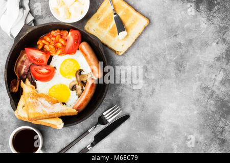 Prima colazione inglese tradizionale - pancetta, salsicce, uova fritte, pomodori, fagioli, funghi, toast con burro - nel recipiente di cottura e caffè sul dorso grigio Foto Stock