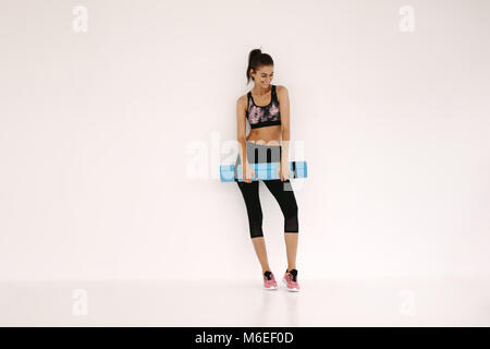 Sportive in piedi con materassino yoga in fitness studio. Un sano modello femminile in piedi su sfondo bianco con materassino yoga dopo allenamento. Foto Stock