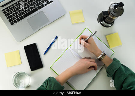 La scrittura nel blocco note al banco con lo smartphone e il computer portatile Foto Stock