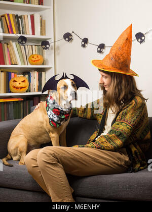 Giovani femmine persona con staffordshire terrier puppy in costume di halloween seduto sul divano nel soggiorno con jack'o'lanterna caqrved zucche in backgroun Foto Stock