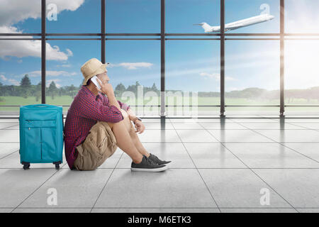 Vista laterale del uomo asiatico seduto e parlando al cellulare mentre si è in attesa sul piano terminal aeroporto Foto Stock