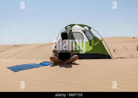 Una donna utilizza e carica il proprio computer portatile con pannelli solari portatili accanto alla tenda da campeggio nel deserto, autosufficiente, eco-compatibile, sostenibile Foto Stock