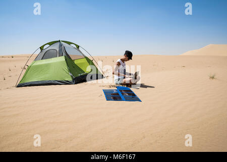 Donna che usa il suo computer portatile per la ricarica con pannelli solari flessibili nel deserto accanto alla sua tenda da campeggio, autosufficienza e sostenibilità in un'area remota Foto Stock