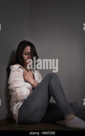 Giovane donna premuto, tirando la sua camicia in modo da coprire se stessa, mentre è seduto sul pavimento, appoggiata contro una parete. Foto Stock