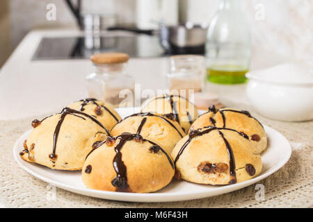 Close-up di immagine sfornati freschi panini a croce. Fatta in Casa dolce di Pasqua il pane sulla tavola servita su piastra bianca nel moderno ambiente cucina Foto Stock