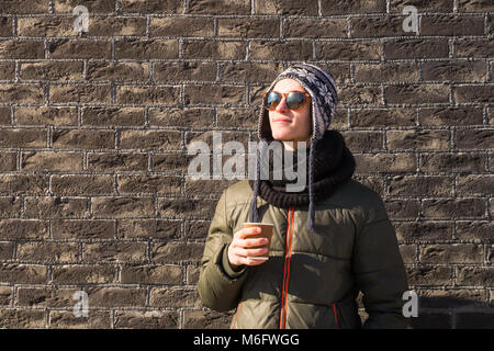 Giovane uomo in abbigliamento invernale tiene tazza di caffè. Bello teenage persona di sesso maschile in occhiali da sole e cappello di maglia bevande bevande calde sul luminoso winter Foto Stock