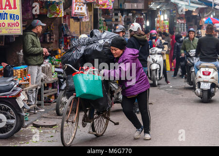 Una donna che spinge la sua bicicletta caricata con pesanti sacchi di colore nero ad Hanoi, Vietnam Foto Stock