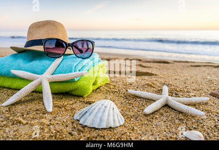 Estate accessori come occhiali da sole, asciugamani, cappello, solare, conchiglie e stella di mare sulla spiaggia di sabbia in Portogallo Foto Stock