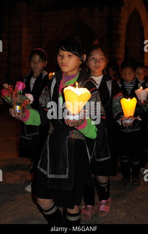 Giovani felici e solenne Hmong ragazze vestiti in abiti tradizionali tenendo candele e fiori celebrare la Vergine Maria a Sapa chiesa di pietra di notte. Foto Stock