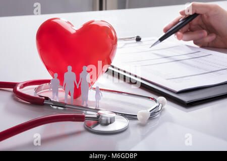 Uno stetoscopio, Famiglia carta tagliato e cuore rosso vicino al medico la mano della relazione di analisi Foto Stock