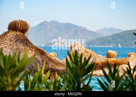 Ombrelloni con paglia in primo piano su una spiaggia soleggiata con un mare cristallino e una catena montuosa sullo sfondo Foto Stock