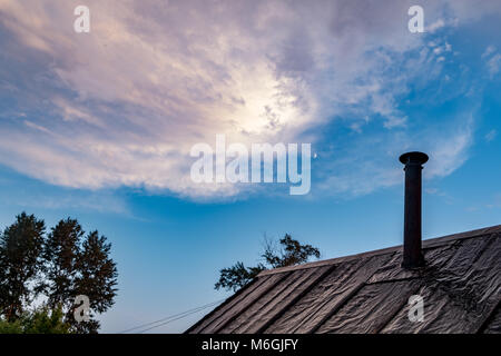 Cielo in prima serata con un mix di sfumature rosa e blu. In primo piano, il tetto usurato con tetti in feltro e camino metallico contrasta con lo sfondo naturale Foto Stock