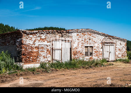 Abbandonata vecchia cowshed di muro di mattoni stagionato e con cancello in legno fatiscente nelle giornate di sole in campagna Foto Stock