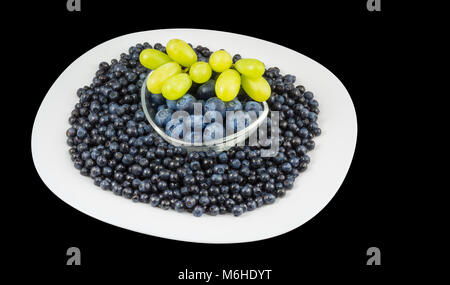 Organici di mirtilli e grappolo di uva. Vaccinium myrtillus. Grandi e piccoli frutti a bacca bianca sulla piastra e ciotola di vetro. Isolato su sfondo nero. Foto Stock