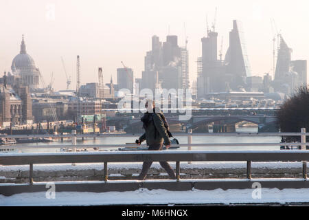 Un " commuter " passeggiate sulla neve coperto Waterloo Bridge con la City di Londra quartieri finanziari di grattacieli, la cattedrale di St Paul e edifici alti Foto Stock