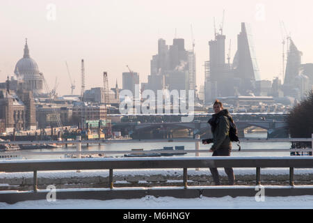 Un " commuter " passeggiate sulla neve coperto Waterloo Bridge con la City di Londra quartieri finanziari di grattacieli, la cattedrale di St Paul e edifici alti Foto Stock