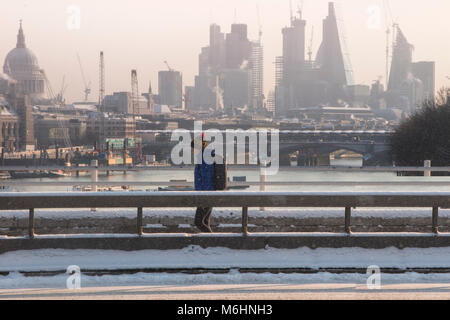 Un " commuter " passeggiate sulla neve coperto Waterloo Bridge con la City di Londra quartieri finanziari di grattacieli, la cattedrale di St Paul e edifici alti Foto Stock