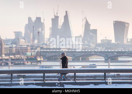 Un " commuter " passeggiate sulla neve coperto Waterloo Bridge con la City di Londra quartieri finanziari di grattacieli, la cattedrale di St Paul e edifici alti Foto Stock