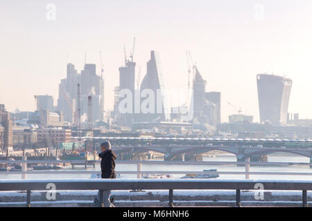 Un " commuter " passeggiate sulla neve coperto Waterloo Bridge con la City di Londra quartieri finanziari di grattacieli, la cattedrale di St Paul e edifici alti Foto Stock