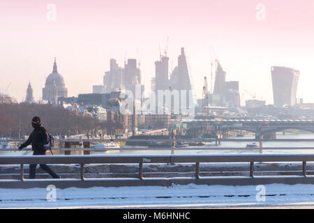 Un " commuter " passeggiate sulla neve coperto Waterloo Bridge con la City di Londra quartieri finanziari di grattacieli, la cattedrale di St Paul e edifici alti Foto Stock