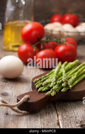 Ingredienti per una sana prima colazione a base di asparagi freschi su un tagliere, pomodori, uova sul vecchio sfondo in legno in stile rustico Foto Stock